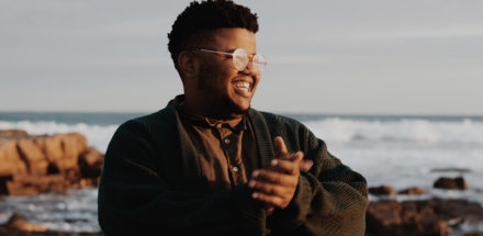 Young man with glasses, rubbing his hands together with the sea and cliffs in the background.