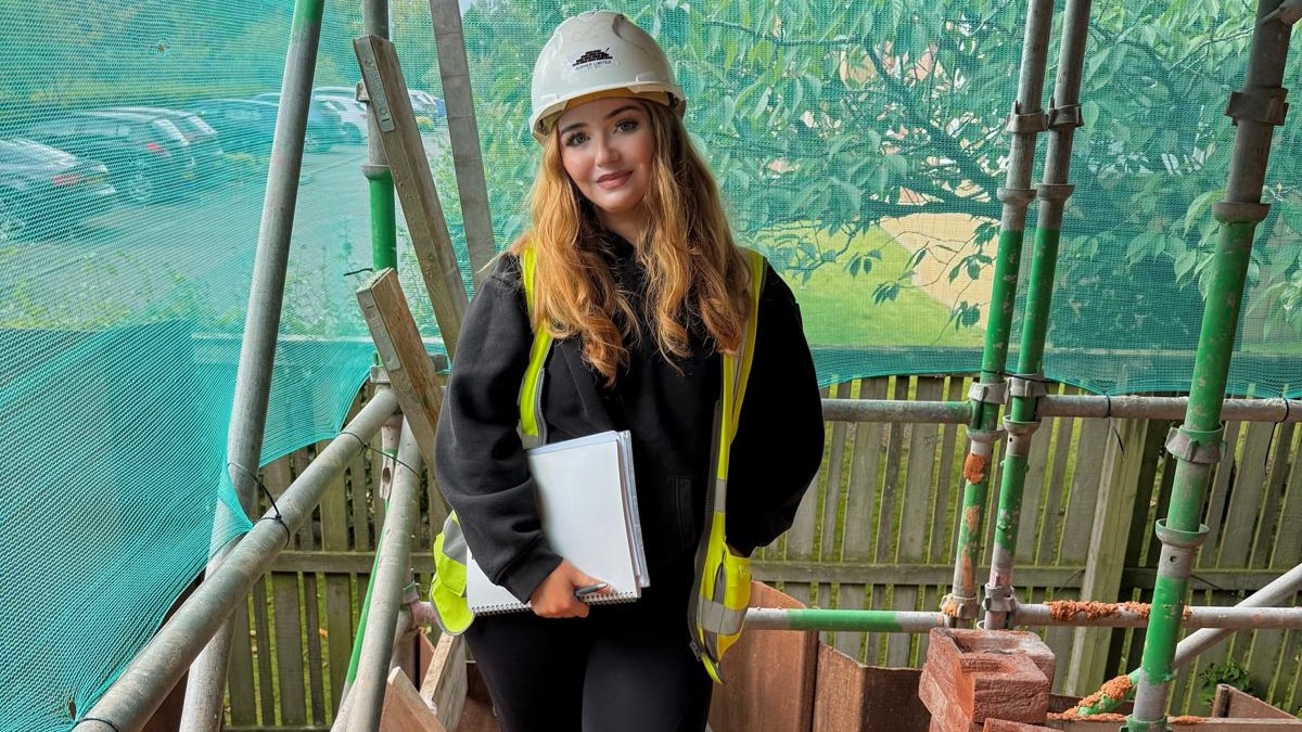 A young woman wearing a hard hat and high-visibility vest stands on scaffolding at a construction site.