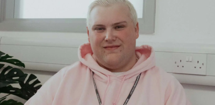 Man with blonde hair wearing a pink hoodie and a lanyard sitting in a classroom.
