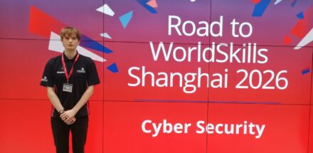 Charlie standing next to a sign which reads 'Road to WorldSkills Shanghai 2026'