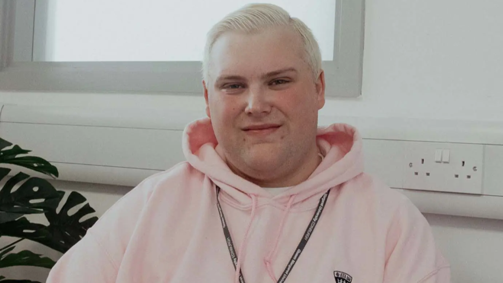 Man with blonde hair wearing a pink hoodie and a lanyard sitting in a classroom.